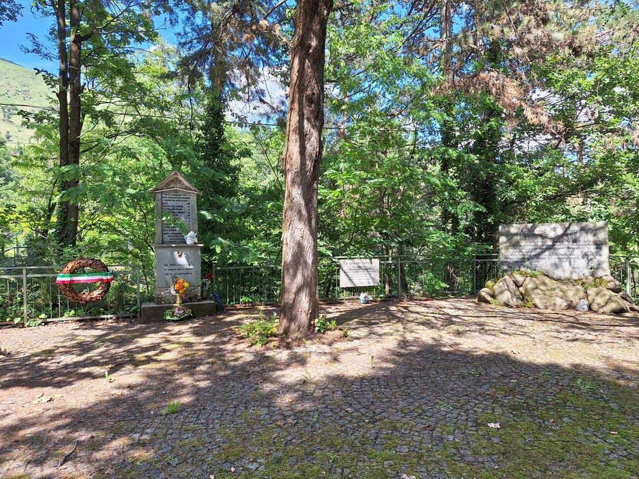 Lapide commemorativa circondata da alberi, con fiori e decorazioni, in un contesto naturale tranquillo.