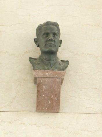 Busto bronzeo di uomo su supporto in marmo, affisso a una parete liscia e chiara.