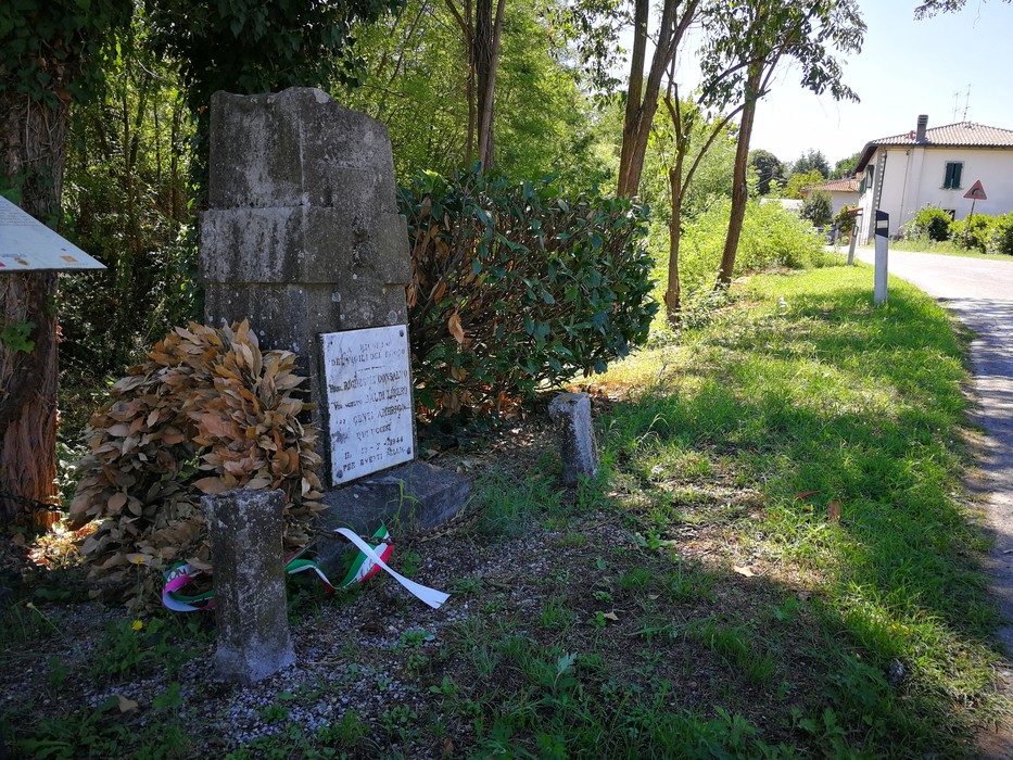 Cippo in pietra con lapide incisa, ai margini della strada tra vegetazione, con fiori e nastro tricolore ai piedi.