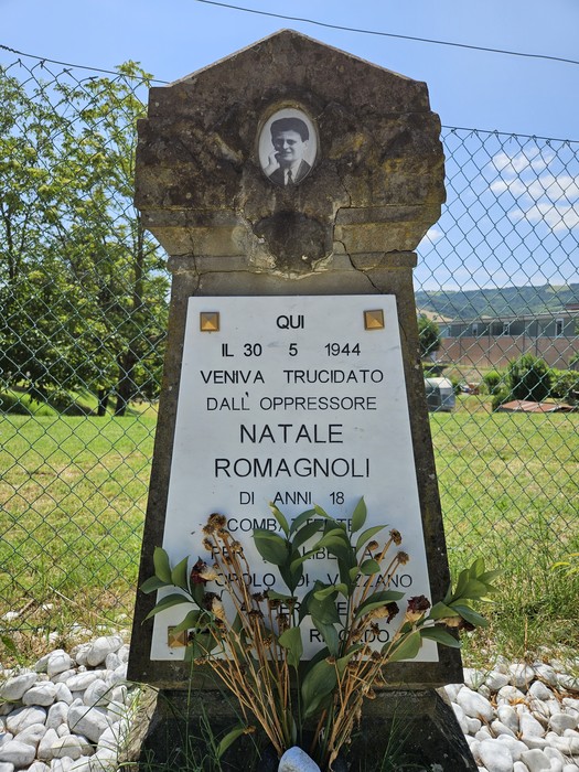 Cippo commemorativo dedicato a Natale Romagnoli, con foto e decorazioni floreali, situato in un'area verde.