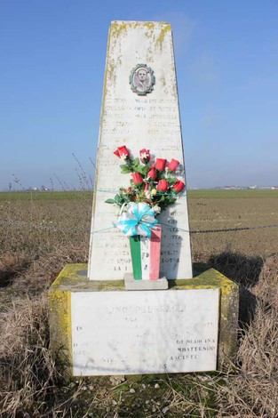 Cippo commemorativo a Pederzoli, ornato con fiori rosso e verde, immerso in un paesaggio rurale.