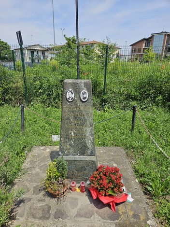 Cippo commemorativo con cerchi fioriti e fotografie, immerso nel verde di un'area urbana.