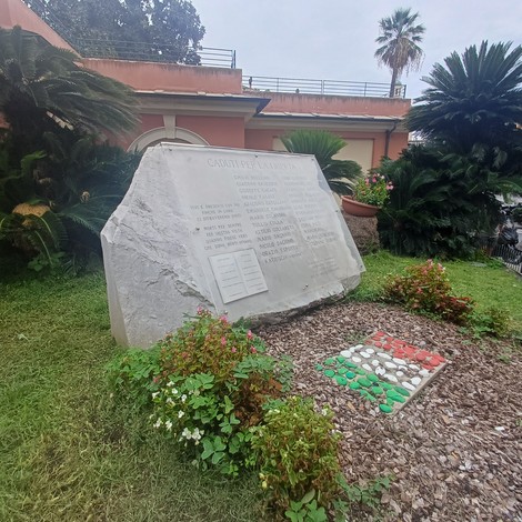 Blocco di pietra grezza con iscrizione, in un giardino con aiuole fiorite e un mosaico tricolore a terra.