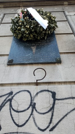 Lapide commemorativa di Rinaldi con ghirlanda di fiori, affissa su una parete con graffiti.