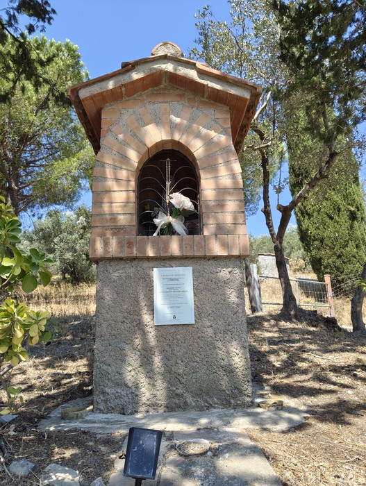 Tabernacolo in mattoni con nicchia ad arco, targa e fiori, tra alberi in un'area verde.