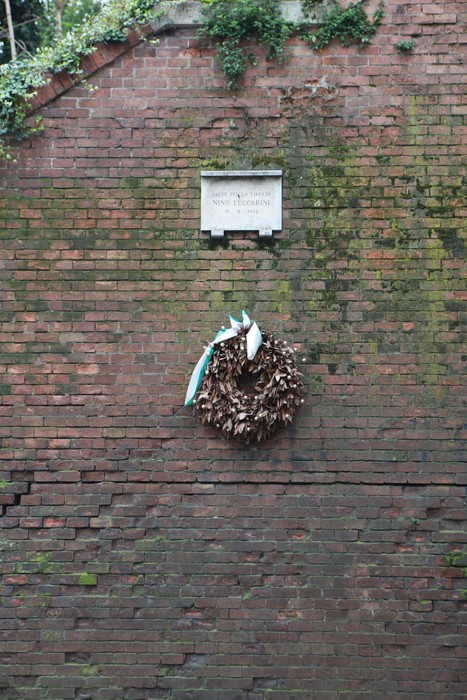 Lapide commemorativa su un muro di mattoni, con una corona di foglie e nastri appesi.