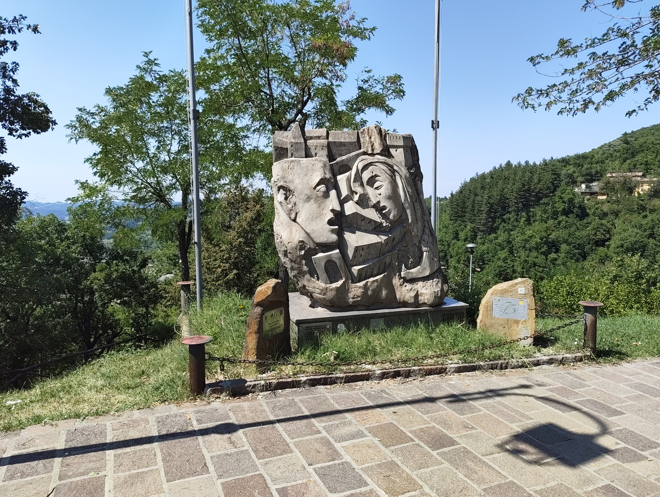 Scultura in pietra con volti stilizzati, su basamento tra alberi lungo un belvedere panoramico.