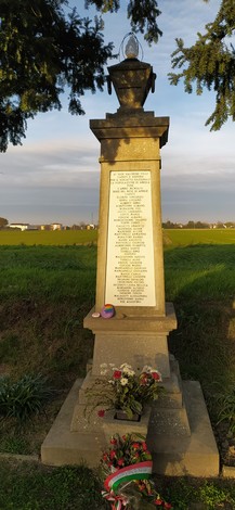 Monumento ai caduti con una targa commemorativa, circondato da fiori e un paesaggio rurale sullo sfondo.