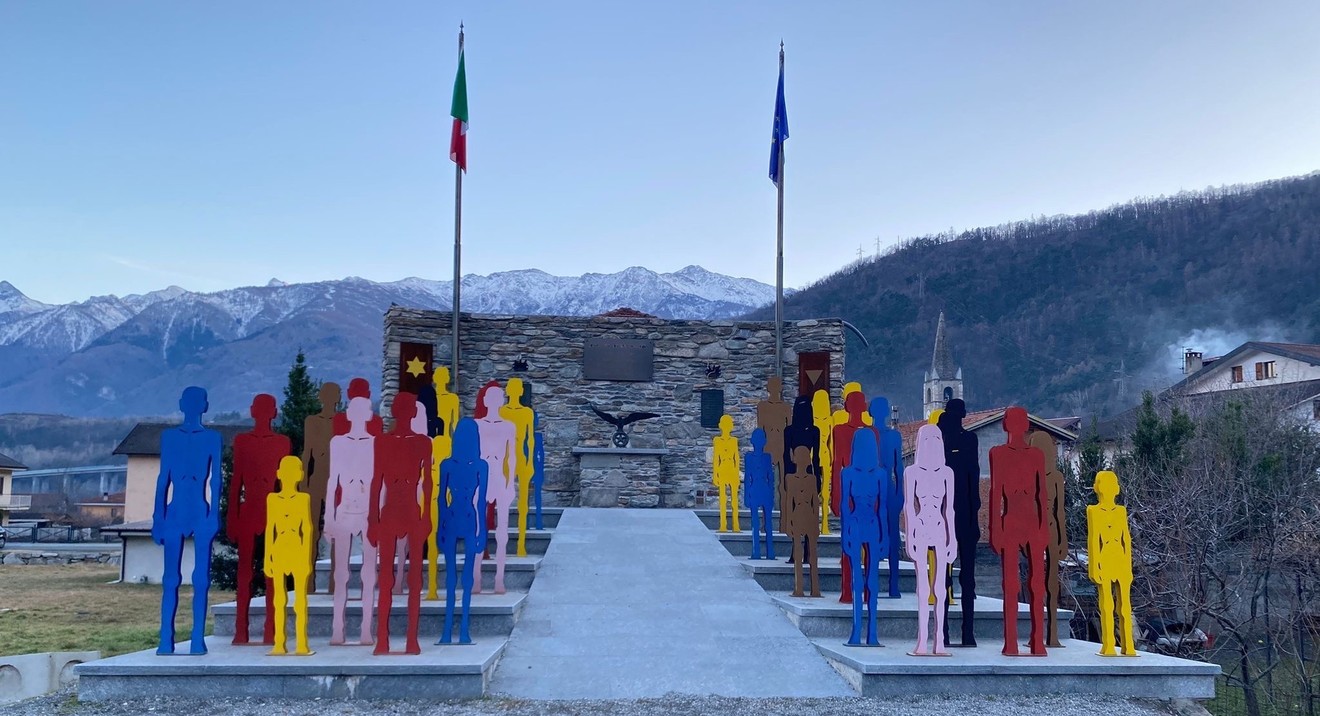 Monumento ai caduti di Venaus, con silhouette colorate e bandiere, sullo sfondo delle montagne.