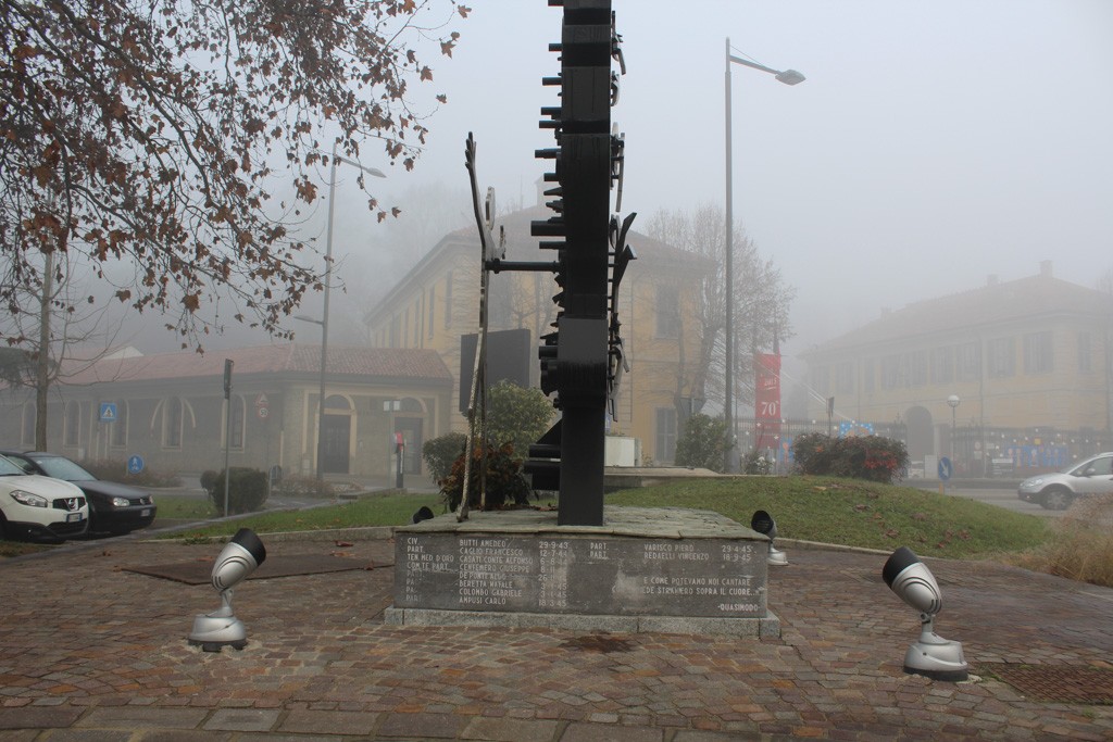 Monumento astratto in metallo scuro su base in pietra con iscrizione, in una piazza avvolta dalla nebbia.
