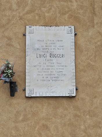 Lapide commemorativa di Luigi Ruggeri, con decorazione floreale e scritte commemorative su sfondo murale.
