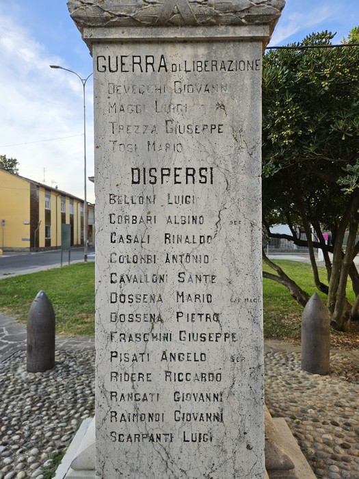 Monumento in pietra con iscrizione dedicata ai caduti, affiancato da due proiettili decorativi in una piazza alberata.