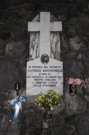 Lapide commemorativa in marmo dedicata a Carmelo Baronchelli, con fiori di omaggio e una croce.