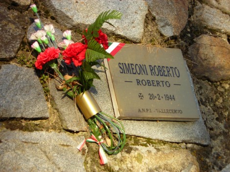 Targa in memoria di Roberto Simeoni, accompagnata da fiori rossi, collocata su una pietra.