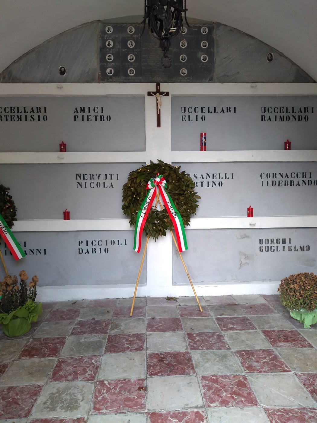 Sacrario con nomi delle vittime, candele e ghirlande commemorative in un ambiente sobrio e rispettoso.