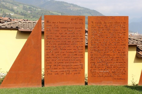 Monumento a Seghezzi, opera in metallo con testi inciso, in un paesaggio montano. Design semplice e contemplativo.