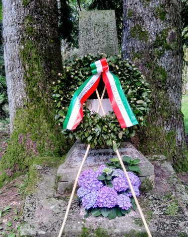 Cippo commemorativo decorato con una corona di foglie e nastro tricolore, circondato da alberi e fiori.