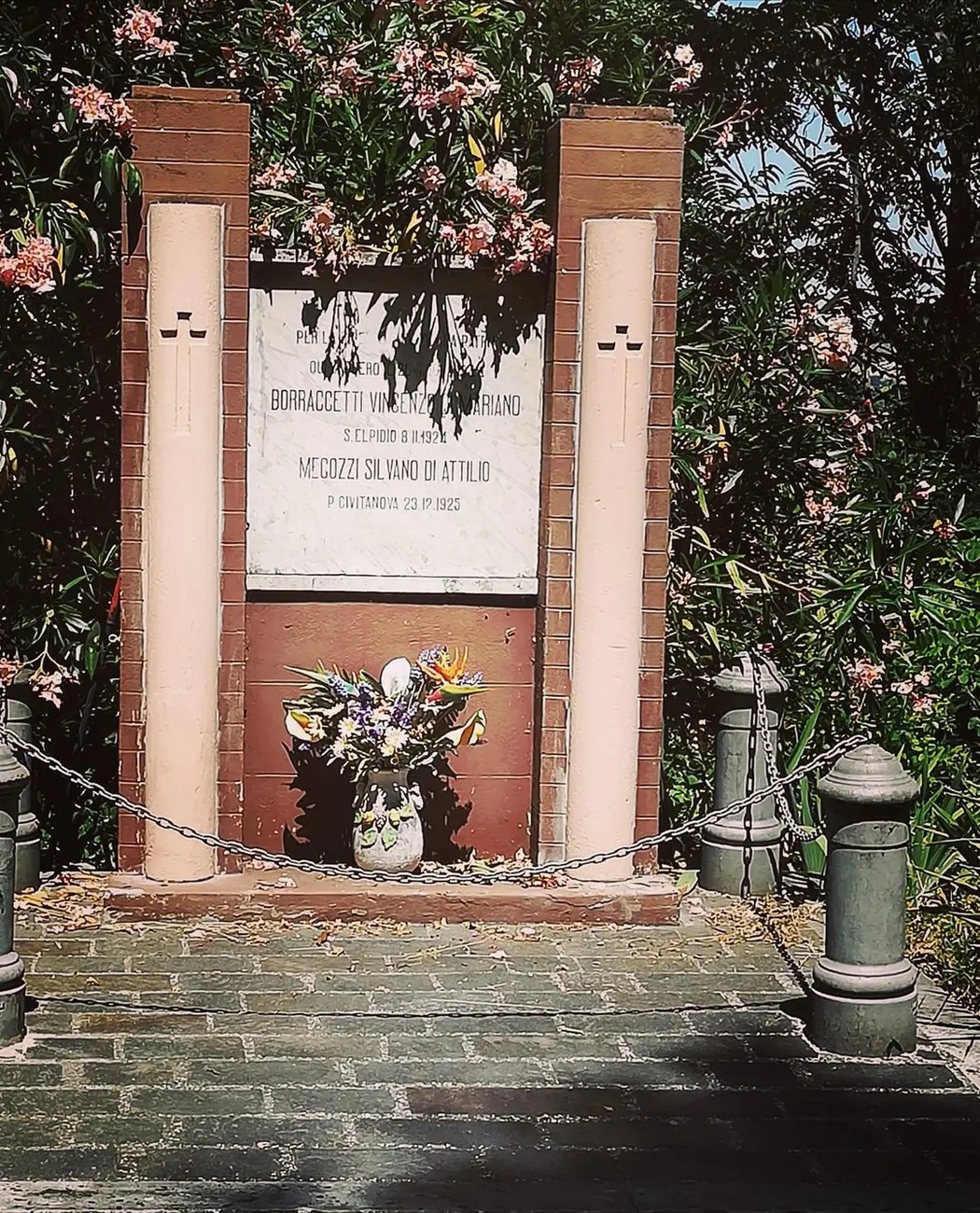 Monumento in muratura con lapide incisa tra due colonne, vaso di fiori davanti e rampicanti fioriti sopra.