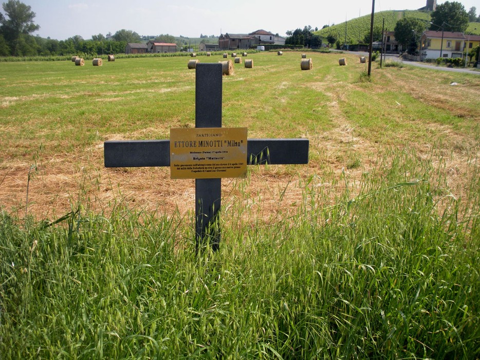 Croce commemorativa di Ettore Minotti in un campo, circondata da un paesaggio rurale.