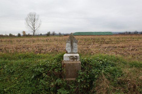 Cippo commemorativo in un campo, con sfondo di cielo nuvoloso e vegetazione circostante.
