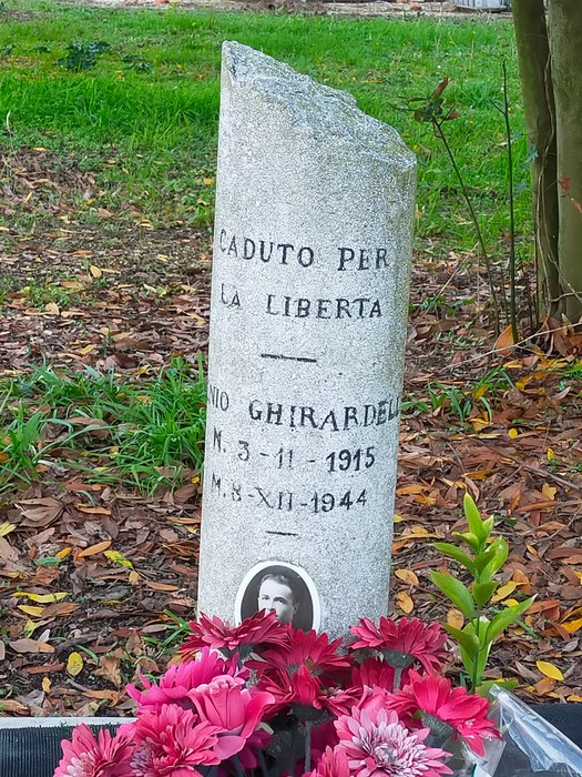 Cippo commemorativo dedicato a Ghirardelli, caduto per la libertà, adornato con fiori e ricordi.