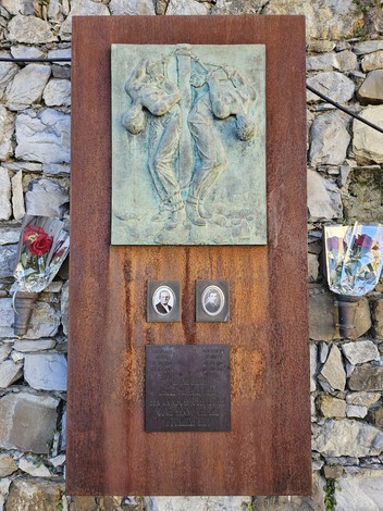Targa commemorativa in metallo su muro di pietra, con bassorilievo e foto di persone, circondata da fiori.