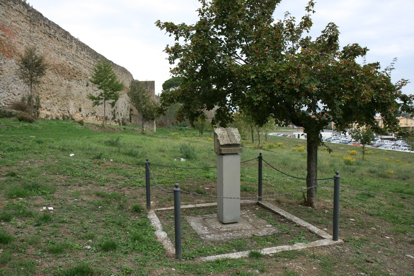 Cippo a Meuret circondato da un prato verde, con alberi e mura storiche sullo sfondo.