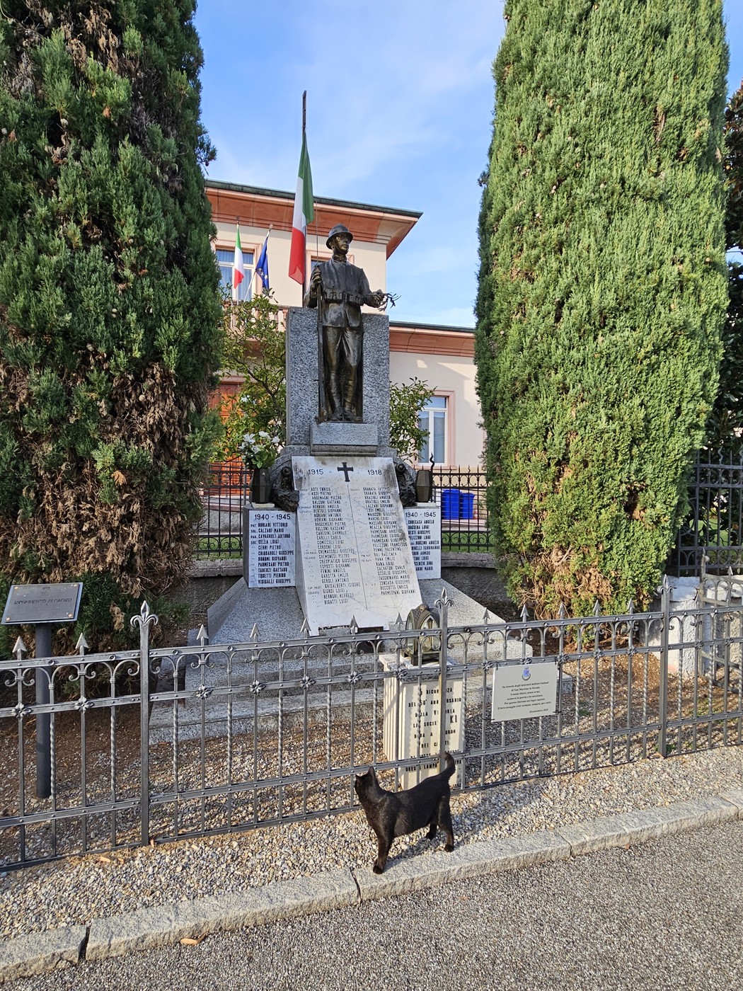 Monumento in pietra con statua bronzea su piedistallo con iscrizione, tra due cipressi, recinzione in ferro.