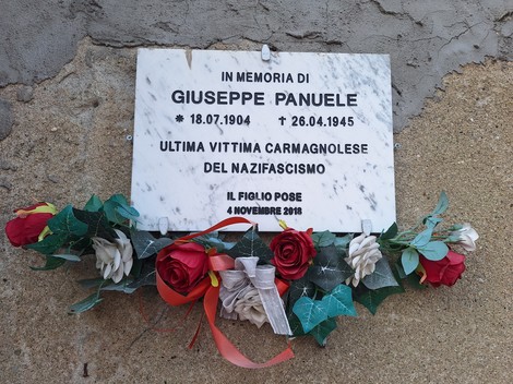 Lapide in marmo su parete intonacata, con iscrizione e corona di fiori artificiali appoggiata sotto.