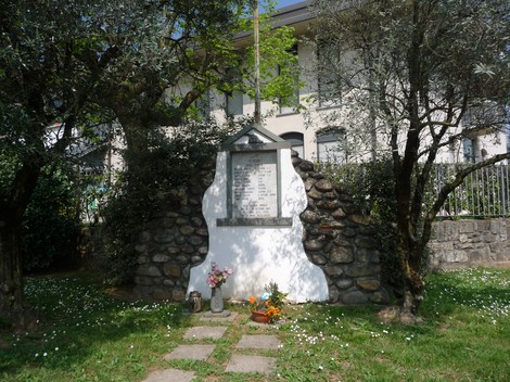 Lapide commemorativa ai caduti partigiani, con decorazioni floreali e un contesto naturale di alberi e prato.