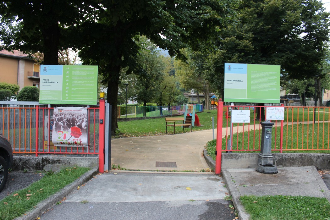 Entrata di un parco con cancelli e pannelli informativi circondati da alberi e aree verdi.