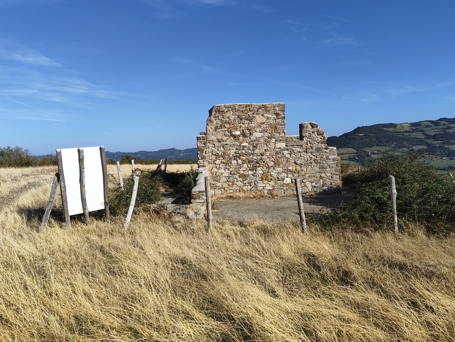 Rovine di un edificio in pietra tra campi recintati, con pannello informativo e colline sullo sfondo.
