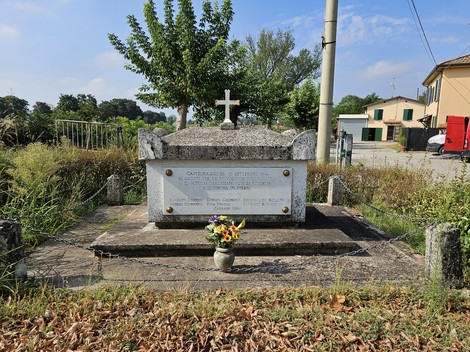 Monumento ai Martiri della Biscia, con decorazioni floreali e una croce, immerso in un paesaggio rurale.