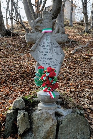 Cippo in pietra a forma di croce con iscrizione, ornato da fiori tricolori, in un bosco su un basamento di sassi.