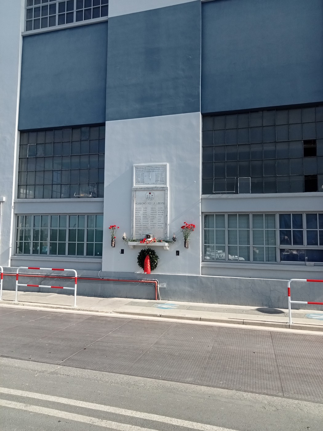 Lapide con iscrizione sulla facciata di un edificio industriale, ornata da fiori rossi e una corona.
