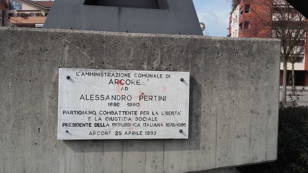 Monumento commemorativo ad Alessandro Pertini con iscrizione su base in pietra, circondato da un'area urbana.