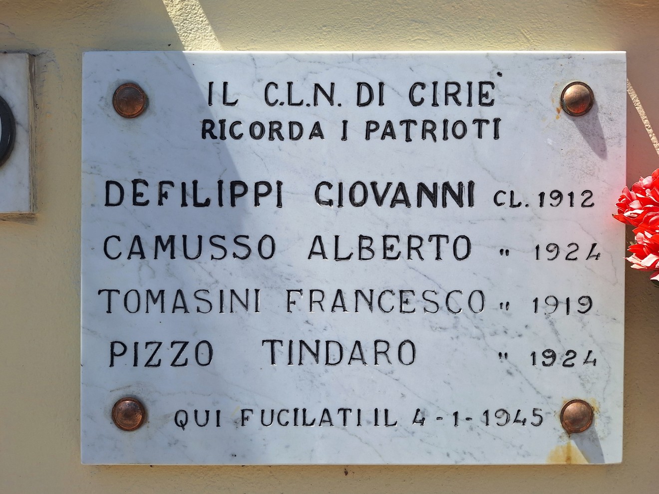 Lapide marmorea con iscrizione a rilievo su parete, fissata con quattro borchie, fiore rosso a lato.