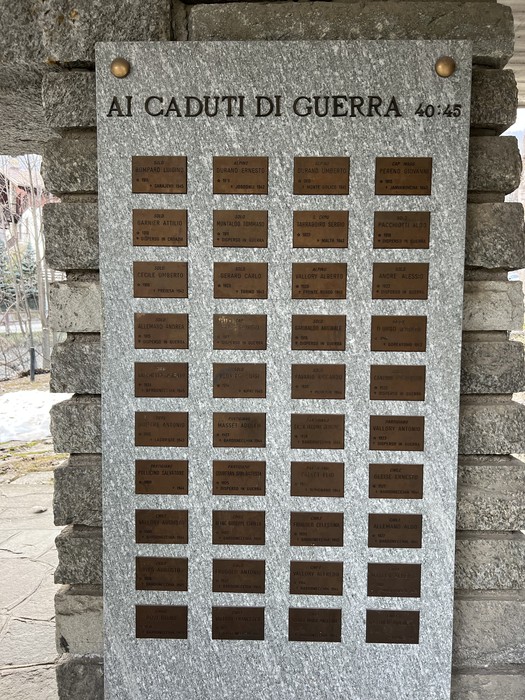 Lapide in marmo con targhe metalliche disposte in file, fissata su parete esterna in pietra.