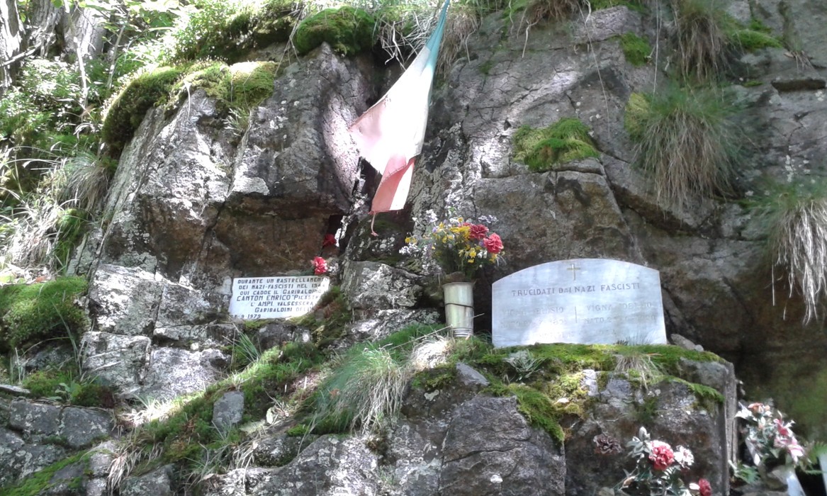 Lapidi commemorative all'Alpe Morcei, con fiori e bandiera tricolore, in un ambiente naturale suggestivo.
