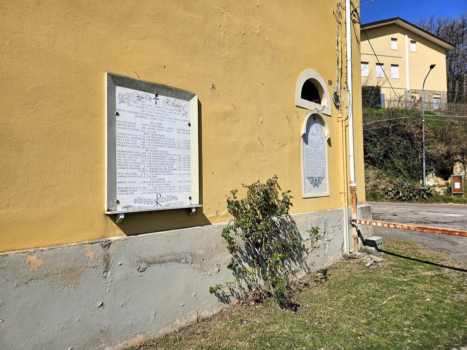 Lapide con elenco di nomi e croce incisa sulla parete gialla di un edificio, accanto a una finestra ad arco.