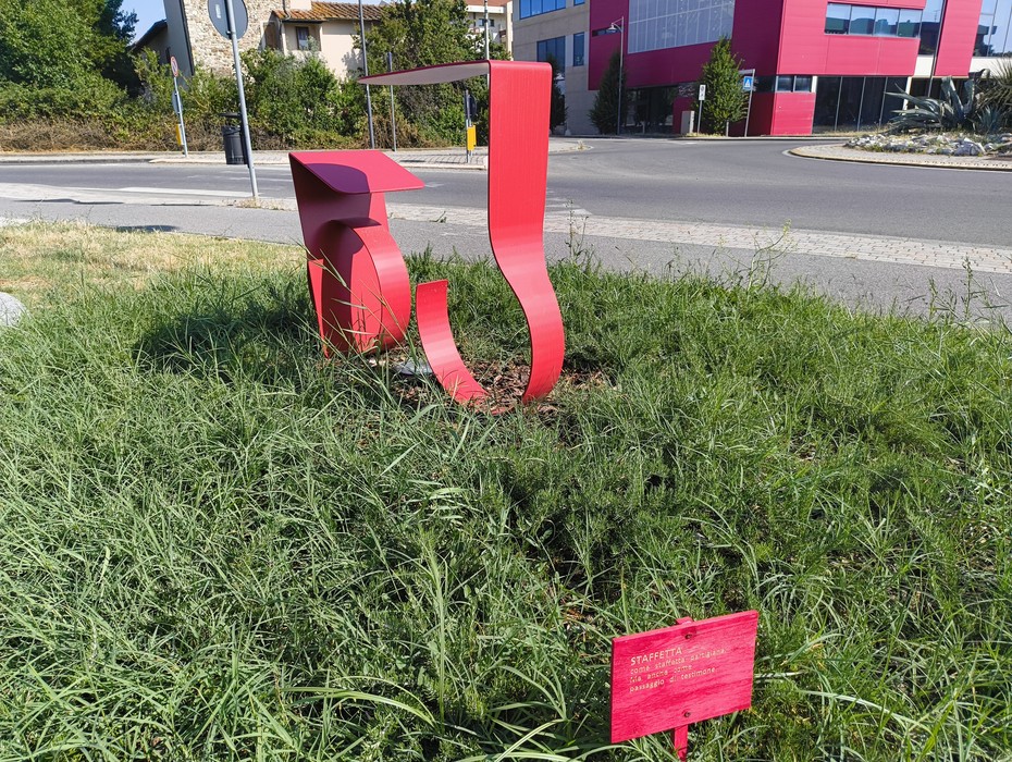 Scultura astratta rossa su prato verde, simbolo di resilienza femminile in un contesto urbano.