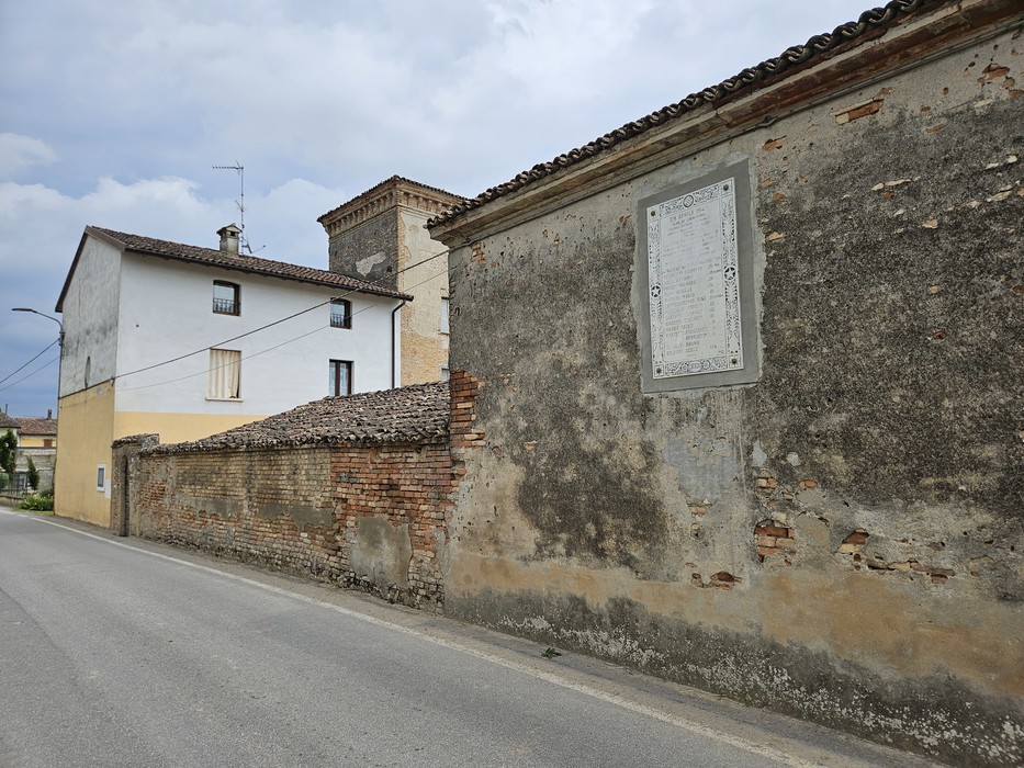 Lapide commemorativa murata su un edificio in una strada di Cavezzo, circondata da un paesaggio urbano.