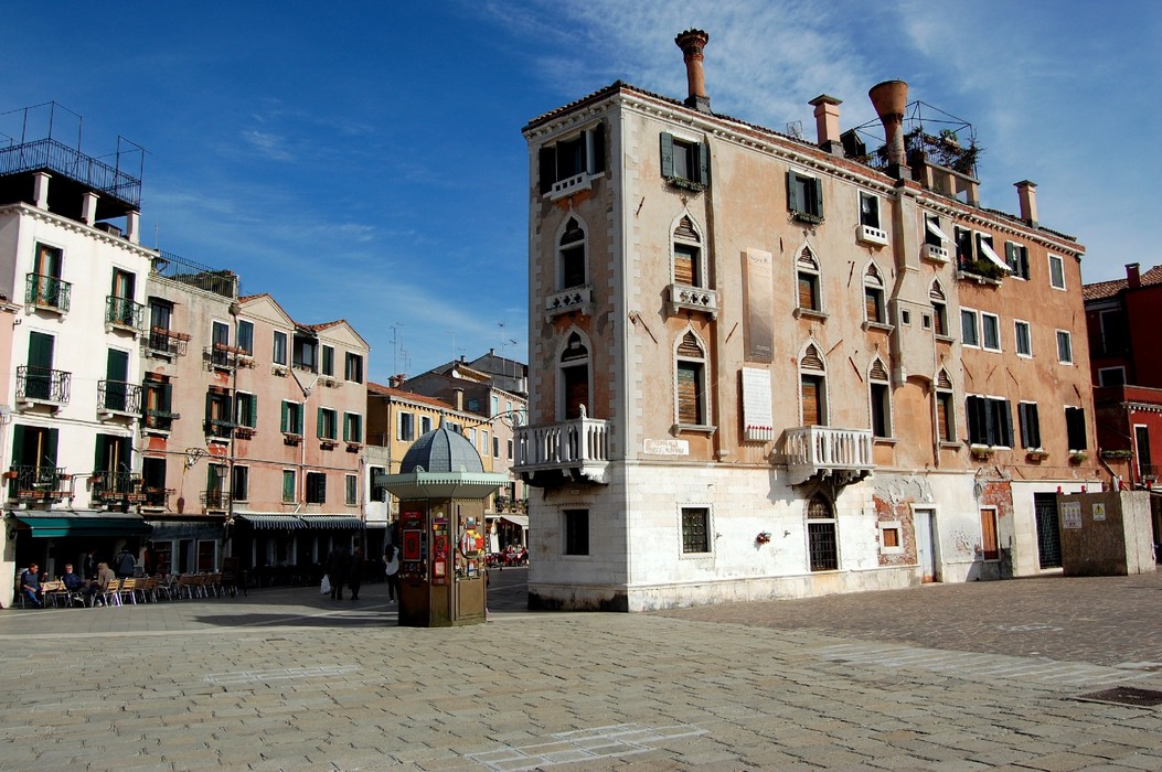 Lapide commemorativa in una piazza storica, circondata da edifici e un cielo azzurro.
