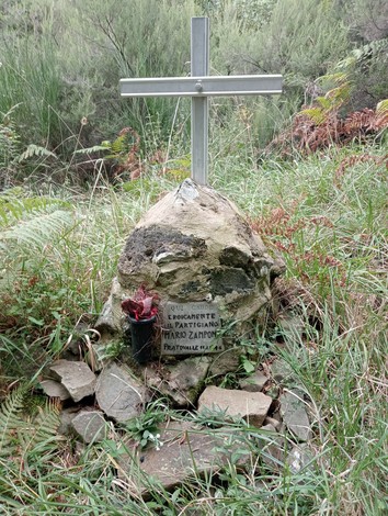 Cippo commemorativo con croce in un'area verde, circondato da piante e rocce, in ricordo di un partigiano.