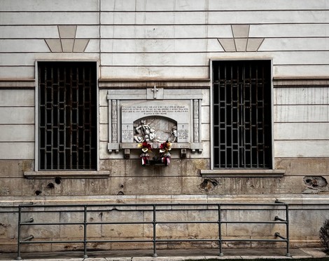 Lapide commemorativa sull'ex palazzo delle poste, con struttura architettonica sobria e fiori deposti.