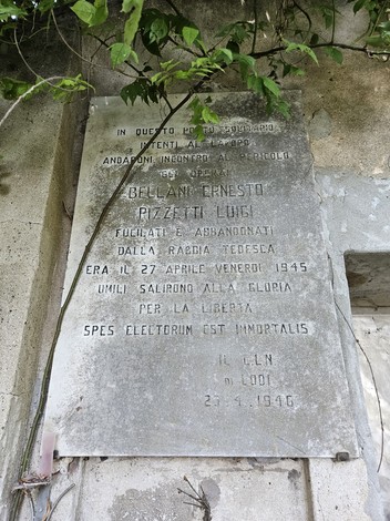 Lapide in marmo con iscrizione, fissata a una parete esterna, incorniciata da rami e foglie.
