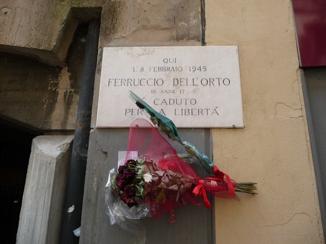 Lapide commemorativa dedicata a Ferruccio Dell'Orto, con fiori, situata su un muro.