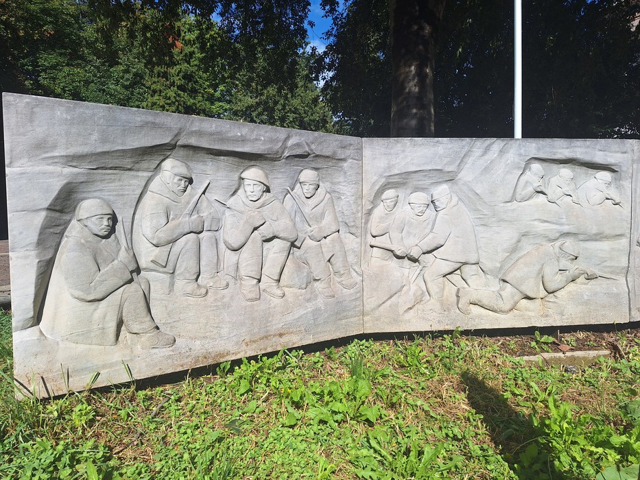Monumento in pietra a due pannelli con bassorilievi di soldati prigionieri, collocato su un prato.