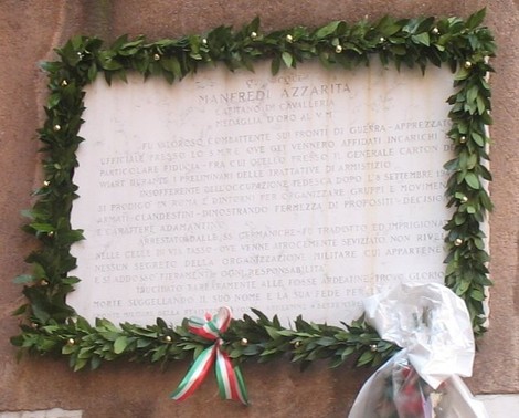 Lapide commemorativa dedicata a Manfredi Azzarita, adornata da una corona di alloro e un mazzo di fiori.