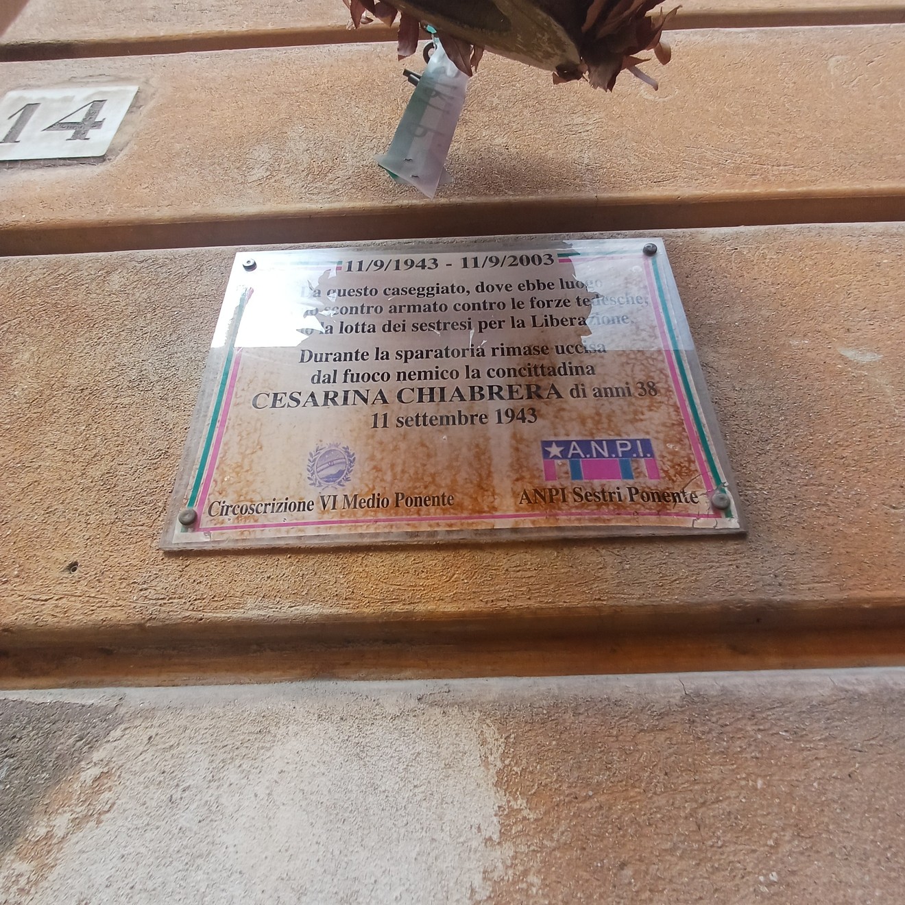 Lapide in marmo con iscrizione, fissata alla parete esterna di un edificio, con fiori appesi sopra.
